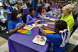  A group of students sit with senior citizens eating snacks, painting and doing crafts